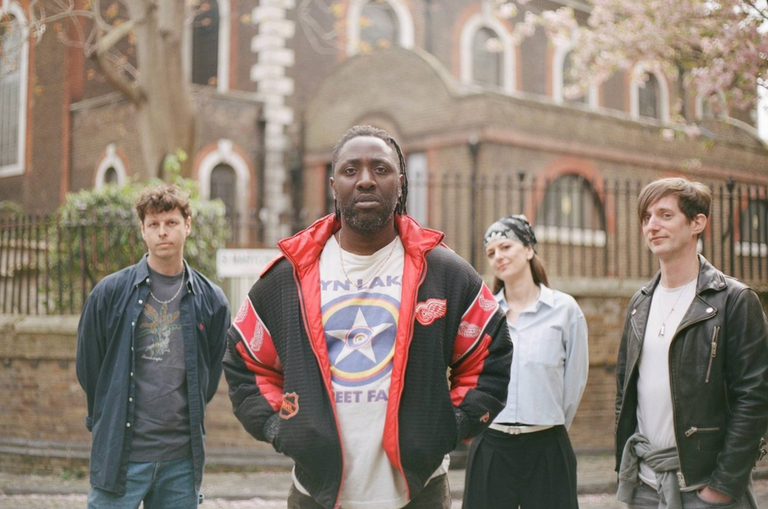 Outdoor group photo of indie rock band Bloc Party standing together in front of a brick building, with frontman Kele Okereke in the center.